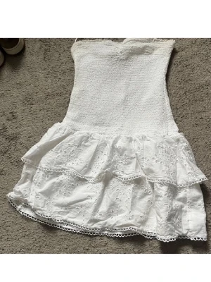 Vit axelbandslös volangklänning - Söt vit axelbandslös klänning med smockad överdel och dubbla volangkjolar i broderad spets. Perfekt för varma dagar och har en romantisk känsla med fina spetsdetaljer och kort längd. Storlek M men passar mig som brukar ha xs 