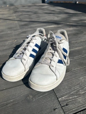 Vita Adidas sneakers med blå ränder - Snygga vita sneakers från Adidas med klassiska blå ränder på sidorna och en röd detalj vid hälen. Skorna har lågt skaft, vita snören och är tillverkade i läder med textilfoder. Perfekta för en sportig och avslappnad stil.
