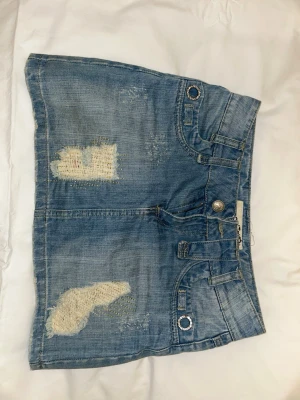 Blå jeanskjol med slitningar - Kort blå jeanskjol från Motivi med råa slitningar och lagade detaljer framtill och baktill. Kjolen har två stora bakfickor med dekorativa knappar och breda bälteshällor. Klassisk femficksmodell i denim med en cool, avslappnad vibe. Passar bra men lite förstor, knappt använd men perfekt till sommaren. ❤️