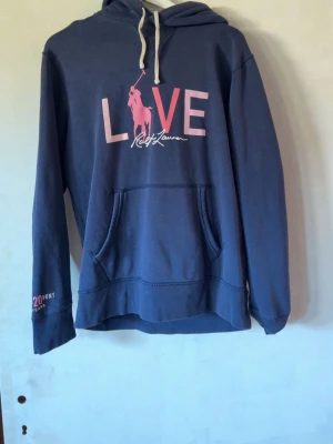 Blå hoodie från Ralph Lauren - Snygg blå hoodie från Ralph Lauren med stor ficka framtill och vit dragsko i huvan. Framsidan har ett rosa och vitt tryck med texten LOVE och den klassiska polospelaren. Mjuk bomullskänsla och avslappnad passform, perfekt för chill dagar.