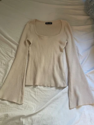 Beige ribbad långärmad topp  - Trendig beige ribbad topp från Gina Tricot med långärmade, utsvängda ärmar och djup rundad ringning. Perfekt för dig som gillar en enkel men ändå snygg stil. Materialet är mjukt och stretchigt, vilket gör toppen bekväm att bära.