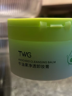 TWG Avocado Cleansing Balm - TWG Avocado Cleansing Balm i en rund burk med ljusgrönt lock och ljusgrön kropp. Perfekt för att ta bort smink och rengöra huden på djupet. Innehåller avokadoextrakt som ger näring och återfuktar huden. Söt avokadoillustration på sidan.