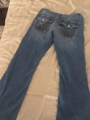 Blå bootcut jeans med broderade fickor - Säljer ett par ljusblå bootcut jeans med snygga broderade bakfickor och knappdetaljer. Jeansen har en klassisk femficksmodell och är tillverkade i denim. Perfekta för dig som gillar detaljer och en avslappnad stil.