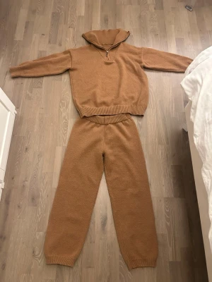 Mysigt brunt stickat loungewear-set - Säljer ett brunt stickat loungewear-set med tröja och byxor. Tröjan har halv dragkedja och hög krage, byxorna har resår i midjan och ribbade muddar. Perfekt för chill dagar hemma. Materialet är mjukt och ger en avslappnad vibe.