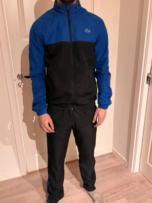 Blå och svart träningsdress Lacoste - Snygg träningsdress från Lacoste i blått och svart. Jackan har dragkedja, hög krage och elastiska muddar. Byxorna har resår i midjan och blåa detaljer längs sidorna. Perfekt för träning eller chill. Materialet är lätt och andas bra. 