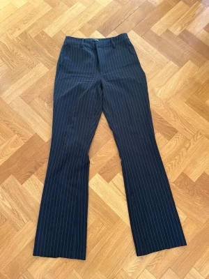 Svarta randiga kostymbyxor - Snygga svarta kostymbyxor med vita tunna kritstrecksränder och utsvängda ben. Klassisk passform med hällor för skärp och diskreta fickor bak. Perfekta för en stilren look. Materialet känns mjukt och följsamt.