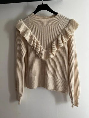 Beige stickad tröja med volang - Trendig beige stickad tröja med ribbad struktur och volangdetalj i V-form framtill men ej baktill. Långa ärmar och rund halsringning. Perfekt för dig som gillar en söt och stilren look. Materialet känns mjukt och bekvämt.