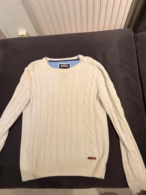 Vit kabelstickad tröja Bondelid - Snygg vit kabelstickad tröja från Bondelid med rund halsringning och diskret läderdetalj vid nederkanten. Tröjan har långa ärmar och klassiskt mönster, perfekt för lager på lager och en clean look.