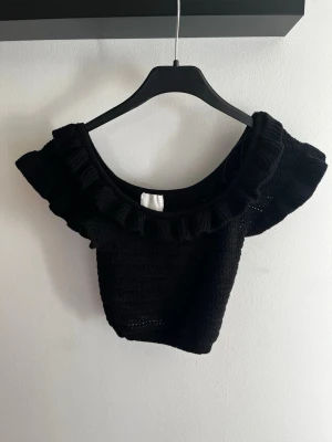 Svart virkad croppad topp H&M XS - Supersöt svart virkad topp från H&M i croppad modell. Toppen har bred volang runt urringningen och är ärmlös, perfekt för en trendig look. Passar dig som gillar detaljer och vill sticka ut lite extra.