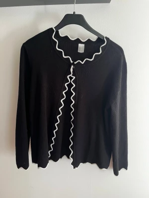 Svart kofta med vågig kant - Snygg svart kofta med långa ärmar och vågiga vita kanter längs halsringning och öppning. Koftan har en öppen design med endast en knapp upptill och är tillverkad i ett mjukt stickat material. Perfekt för att addera en cool detalj till din outfit. Köpt begagnad men ej använd av mig.