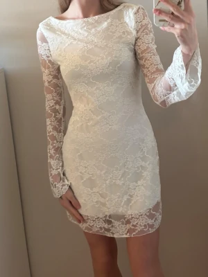 Vit spetsklänning med lång ärm - Säljer en elegant vit kortklänning med långärmade spetsdetaljer och figurnära passform. Klänningen har transparenta spetsärmar och ett vackert blommigt mönster över hela tyget. Perfekt för dig som vill ha en romantisk och stilren look.