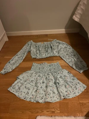 Ljusblått blommigt set med volanger - Säljer ett ljusblått tvådelat set med blommönster. Toppen har långa puffärmar och resår vid axlar och midja. Kjolen har bred smock i midjan och dubbla volanger. Perfekt för en söt och somrig look!