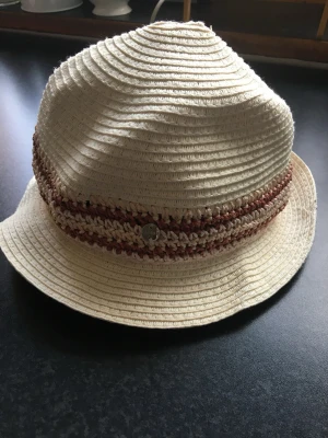 Beige stråhatt med brun detalj - Snygg beige stråhatt med rundad form och bredare brätte. Hatten har ett flätat band i beige och brunt runt kronan samt en liten metallknapp som dekoration. Perfekt för soliga dagar och ger en chill vibe till din outfit.