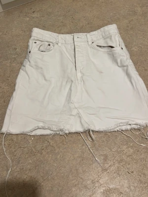 Vit jeanskjol med rå kant - Vit jeanskjol med femficksmodell och rå, fransig kant nertill. Kjolen har knapp- och dragkedjestängning framtill och klassiska nitar vid fickorna. Perfekt för en avslappnad och trendig look.