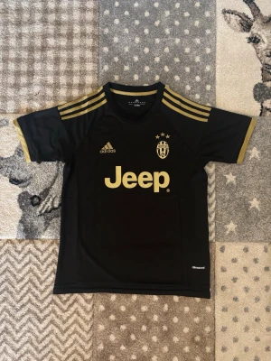 Svart Juventus tröja från Adidas - Säljer en svart Juventus matchtröja från Adidas med guldfärgade detaljer och loggor. Tröjan har korta ärmar, rund hals och är tillverkad i lätt och ventilerande ClimaCool-material. Perfekt för fotboll eller träning.