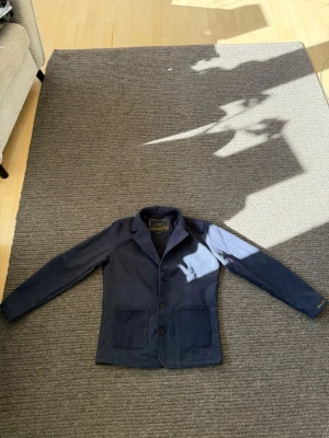 Mörkblå kavaj från Dance De La Sence - Nypris 1800kr. Jag har inte använt denna en ända gång. Stilren mörkblå kavaj med två stora framfickor och två knappar framtill. Klassisk krage och diskret broderad text på ärmslutet. Perfekt för en dressad look. Materialet känns mjukt och följsamt.