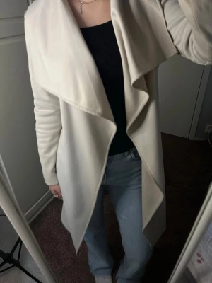 Beige kappa med skärp och vattenfallsärm - Stilren beige kappa med vattenfallsärm och knytskärp i midjan. Kappan har en mjuk, slät yta och faller snyggt längs kroppen. Perfekt för dig som gillar minimalistisk och trendig look. Passar till jeans eller klänning för en cool vardagsstil.