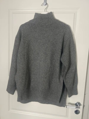 Grå stickad polotröja - Mysig grå stickad polotröja med hög krage och ribbade muddar. Tröjan har en avslappnad passform och är perfekt för kyliga dagar. Materialet ser ut att vara ull eller en ullmix, vilket gör den extra varm och skön.