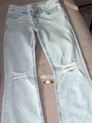 Ljusblå slitna jeans från Nelly - Säljer ett par ljusblå jeans från Nelly med utsvängda ben och slitningar vid knäna. Jeansen har klassisk femficksmodell och coola detaljer med rosa broderi på bakfickorna. Tillverkade i denim med en avslappnad vibe.