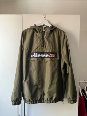 Olivgrön vindjacka från Ellesse - Snygg olivgrön vindjacka från Ellesse med stor logga på bröstet. Jackan har huva med dragsko, halv dragkedja framtill och elastiska muddar. Praktisk magficka och meshfoder på insidan för extra komfort. Perfekt för blåsiga dagar.
