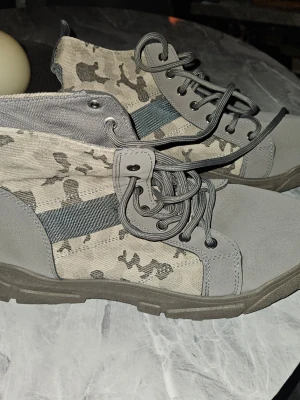 Grå kamouflagekängor Mainalun - Snygga grå kängor från Mainalun med coolt kamouflagemönster och robust sula. Skorna har högt skaft, grå snörning och detaljer i textil och syntetmaterial. Perfekta för dig som gillar en streetstyle-look och vill sticka ut.