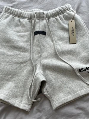 Gråa Essentials shorts med snörning - Säljer ett par ljusgrå shorts från Essentials med snörning i midjan och elastisk resår. Shortsen har en avslappnad passform och logga på benet. Perfekta för sommaren eller chill dagar.