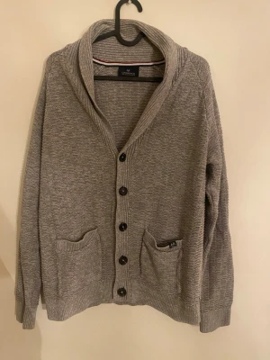 Lindbergh grå cardigan – premiumkvalitet - Snygg grå cardigan från Lindbergh i grovstickad kvalitet med stora knappar framtill och två praktiska fickor. Bred, ribbad krage och ribbade muddar vid ärmslut och nederkant som ger en riktigt clean passform. Perfekt för lager på lager, vardag eller en mer uppklädd stil. Står som storlek L men sitter som en S. 