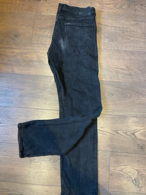 Svarta Lee jeans med rak passform - Säljer ett par svarta jeans från Lee med klassisk rak passform. Jeansen har fem fickor, bälteshällor och en Lee-logga på bakfickan samt läderpatch i midjan. Materialet är mjukt denim i bomull och färgen är snyggt tvättad svart.