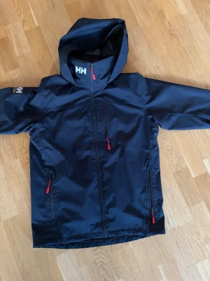 Mörkblå Helly Hansen Jacka W Crew Hooded Mid Plus - Säljer min Helly Hansen jacka i jättefint skick. Den är stilren bekväm och passar till allt både till vardags och när det är kallt ute. Jackan är varm men ändå lätt att ha på sig och känns riktigt bra i kvalitet. Perfekt nu när det börjar bli kallare och man vill ha något som både ser bra ut och håller värmen.  Storlek S.  Jag har använt den mindre än 5 gånger så den är nästan som ny. Den har mest hängt i garderoben och inte kommit till användning.  Ny pris 2000 mitt pris 950