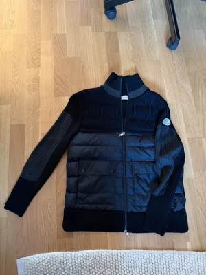 Svart cardiganjacka från Moncler - Snygg svart cardiganjacka från Moncler med quiltad front och stickade ärmar. Jackan har dragkedja, två framfickor med lock och Moncler-logga på ärmen. Materialmix av dun och ull ger både värme och stil. Perfekt till höst och vinter.