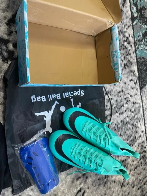 Turkosa fotbollsskor med svart detalj - Säljer ett par turkosa fotbollsskor med svart detalj på tån och snörning framtill. Skorna har en modern, strömlinjeformad design och levereras med ett par blå benskydd samt en svart specialpåse. Perfekt för dig som vill sticka ut på planen.