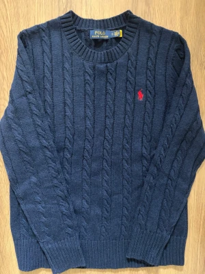 Ralph Lauren Kabelstickad Tröja - Säljer denna kabelstickad mörkblå tröjan från Polo Ralph Lauren med rund halsringning och ribbade muddar. Klassisk röd logga broderad på bröstet. Perfekt för dig som gillar tidlös och stilren design. Materialet känns mjukt och håller värmen.               Tröjan är storlek S och är aldrig använd!                Skriv om ni har några funderingar!😄
