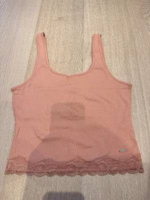 Rosa ribbad linnetopp från Hollister - Supersöt ljusrosa ribbad linnetopp från Hollister med spetskant nertill. Perfekt för sommaren och enkel att styla med jeans eller kjol. Tillverkad i mjuk bomullsmix och har breda axelband för en bekväm passform.