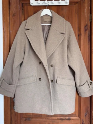 Beige ullkappa från Soyaconcept - Stilren beige ullkappa från Soyaconcept med dubbelknäppning och breda slag. Dekorativa knappar vid ärmsluten och ett diskret prickigt innerfoder. Perfekt för kyliga dagar och ger en klassisk look. Har använt den 1 gång, sen dess har den hängt i garderoben. Köpte den för 1199:- nypris 💘🌸