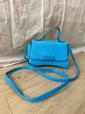 Blå Primark handväska med axelrem - Ny pris 300 (pris kan förhandlas). Snygg liten Primark handväska i blått konstläder med fin dekorativ detalj framtill. Väskan har ett topphandtag och ett långt axelband så du kan bära den både över axeln och i handen. Perfekt för dig som gillar färg och som vill göra outfiten lite mer färgglad.