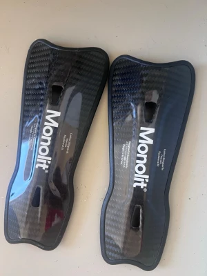Monolit benskydd i kolfiber - Säljer ett par svarta benskydd från Monolit i kolfiber med ergonomisk form och vadderad insida. Lätta och slitstarka, perfekta för dig som vill ha extra skydd och komfort på planen. Snygg design med Monolit-logga på framsidan. 40 gram 
