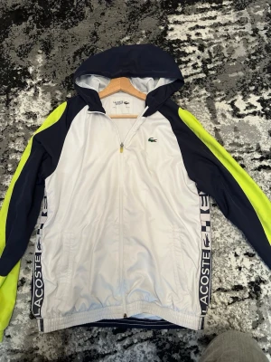 Lacoste vindjacka med huva - Sportig vindjacka från Lacoste med huva, vit front och rygg, mörkblå ärmar och detaljer samt neongula inslag längs ärmarna. Dragkedja framtill, sidofickor och Lacoste-logga på bröstet och längs sidorna. Tillverkad i lätt syntetmaterial.