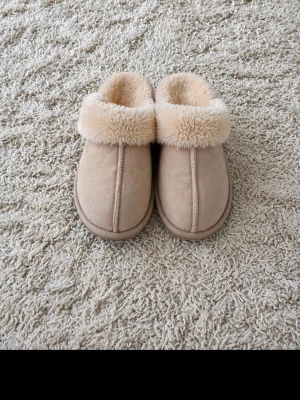 Beige fluffiga tofflor - Mysiga beige tofflor med mjuk fuskpäls runt kanten och insidan. Utsidan är i mockaimitation och sulan är platt. Perfekta för att hålla fötterna varma hemma. Klassisk slip-in modell med rundad tå och enkel design.