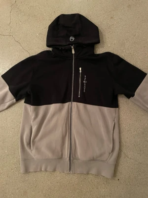 Svart och grå hoodie Sail Racing - Snygg hoodie från Sail Racing i svart och grått med dragkedja framtill och en bröstficka med logga. Hoodien har en stor huva och mjukt foder på insidan. Perfekt för dig som gillar streetwear och vill ha något bekvämt och stilrent.