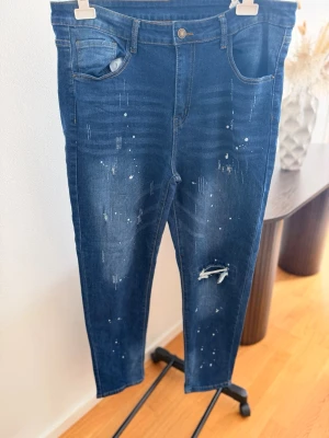 Mörkblå jeans med slitningar - Snygga mörkblå jeans med slitningar och färgstänk för en trendig look. Jeansen har klassisk femficksmodell, normal passform och är tillverkade i bomull. Perfekta för dig som gillar en avslappnad och cool stil.