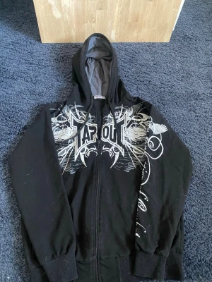 Svart Tapout hoodie med tryck - Cool svart hoodie från Tapout med stort vitt och grått grafiskt tryck över bröstet och ärmen. Hoodien har dragkedja, ribbade muddar och en fodrad huva med snören. Perfekt för dig som gillar streetwear och MMA-inspirerad stil. Hör av er vid frågor eller funderingar 