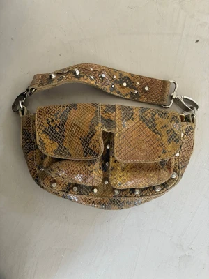  ormmönstrad handväska med nitar - As cool handväska i ormmönster med två framfickor och silverfärgade nitar. Väskan har bred axelrem med nitar och dragkedja upptill. Jättenice som clutch eller som enkel handväska. 