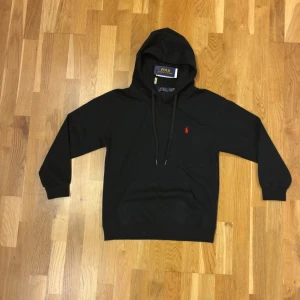 Svart hoodie från Polo Ralph Lauren - Svart hoodie i nyskick från Polo Ralph Lauren med klassisk röd broderad logga på bröstet. Tröjan har huva med snörning, känguruficka och ribbade muddar. Säljer pga fel storlek