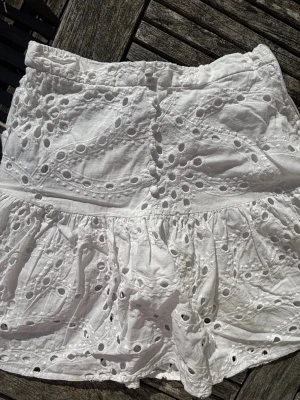 Vit kort kjol med broderade detaljer - Söt vit kortkjol med broderade hålmönster och volang. Kjolen har resår i midjan för skön passform och är tillverkad i ett lätt bomullstyg, perfekt för varma dagar. Den romantiska stilen och de fina detaljerna gör den superfin till sommaren.-kan prissänka,skriv om frågor/bilder, köpt på new yorker men märket är amish