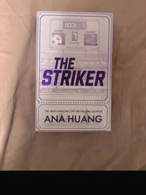 The Striker av Ana Huang - En skönlitterär bok med titeln The Striker av Ana Huang. Omslaget är ljust med lila och svarta detaljer, och boken har sporttema med fotbollsmotiv. Perfekt för dig som gillar romantik och sportiga berättelser.på engelska, har en bruten spine 