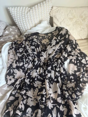 Blommig svart och beige kortklänning - Snygg kortklänning med svart botten och beige blommönster. Klänningen har v-ringning, långärmade transparenta ärmar och smock i midjan för en skön passform. Tillverkad i ett lätt och luftigt material som ger en flowy känsla.