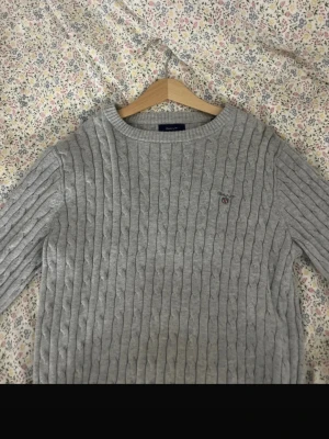Grå kabelstickad tröja från Gant - Säljer en grå kabelstickad tröja från Gant med rund halsringning och diskret broderad logga på bröstet. Tröjan har långa ärmar och ribbade muddar. Perfekt för dig som gillar klassisk och stilren design.