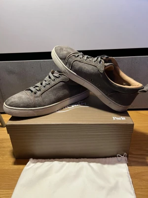 Grå sneakers från Christian Louboutin  - Snygga grå sneakers i mocka med vita sulor och grå skosnören. Klassisk låg modell med diskreta detaljer och bekväm innersula. Perfekta för dig som gillar stilrena och tidlösa sneakers.