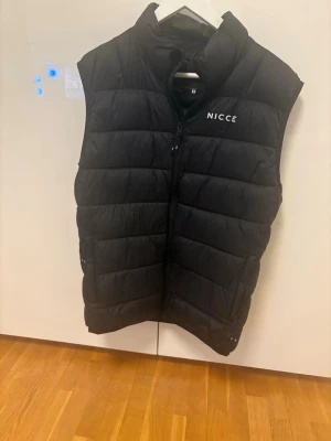Svart dunväst från NICCE - Snygg svart dunväst från NICCE med diskret logga på bröstet. Västen har hög krage, dragkedja framtill och två sidofickor med dragkedja. Perfekt för lager-på-lager och ger en clean streetwear-look.