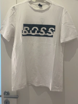 Vit BOSS t-shirt med tryck - Snygg vit t-shirt från BOSS med ett stort, grafiskt tryck i blått och vitt över bröstet. Klassisk rund hals och korta ärmar. Perfekt för en clean och modern stil.